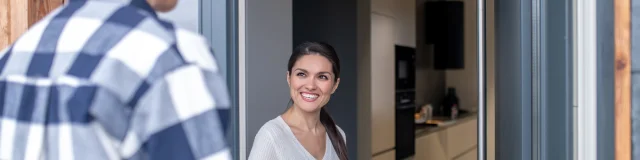 Showing of housing. Young adult smiling woman realtor with folder near open door inviting to house man in plaid shirt standing with his back to camera outdoors